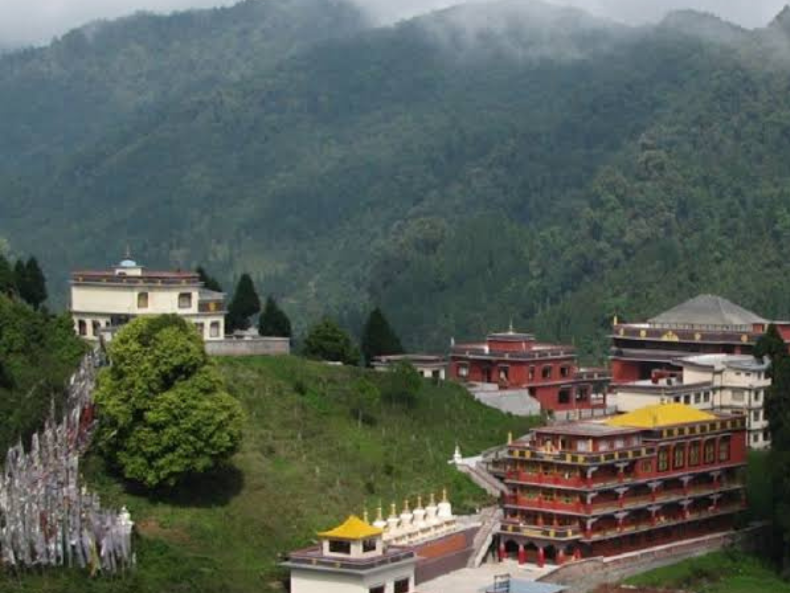 The red and gold monastery at Lava on a hillside with prayer flags and cloud on the ridge behind